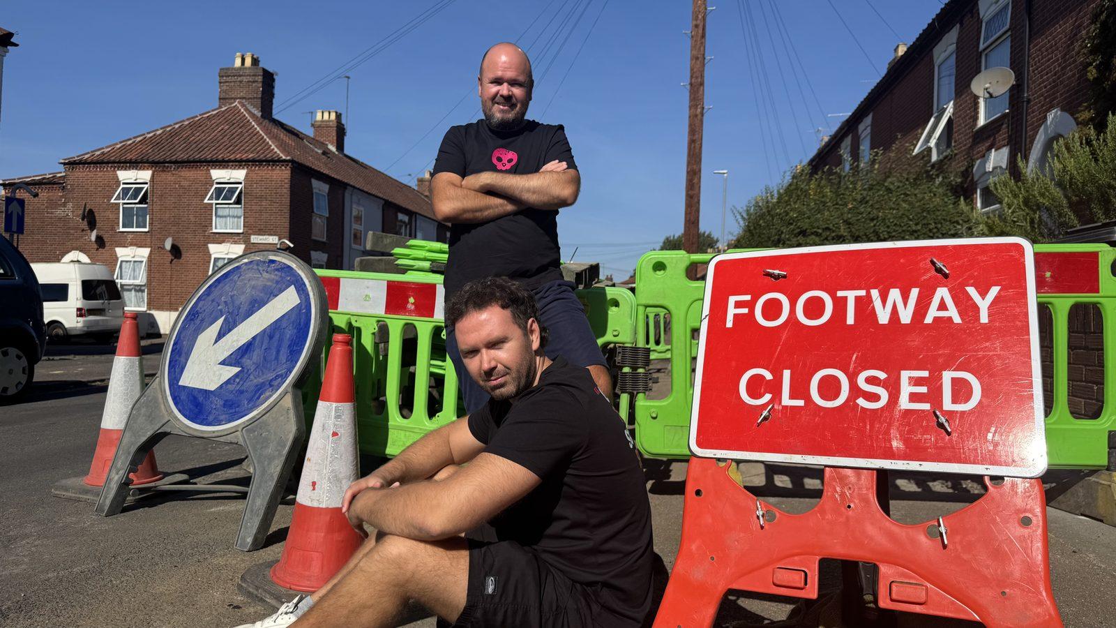 The co-founders beside roadworks barriers and a red “Footway Closed” sign on a residential street.