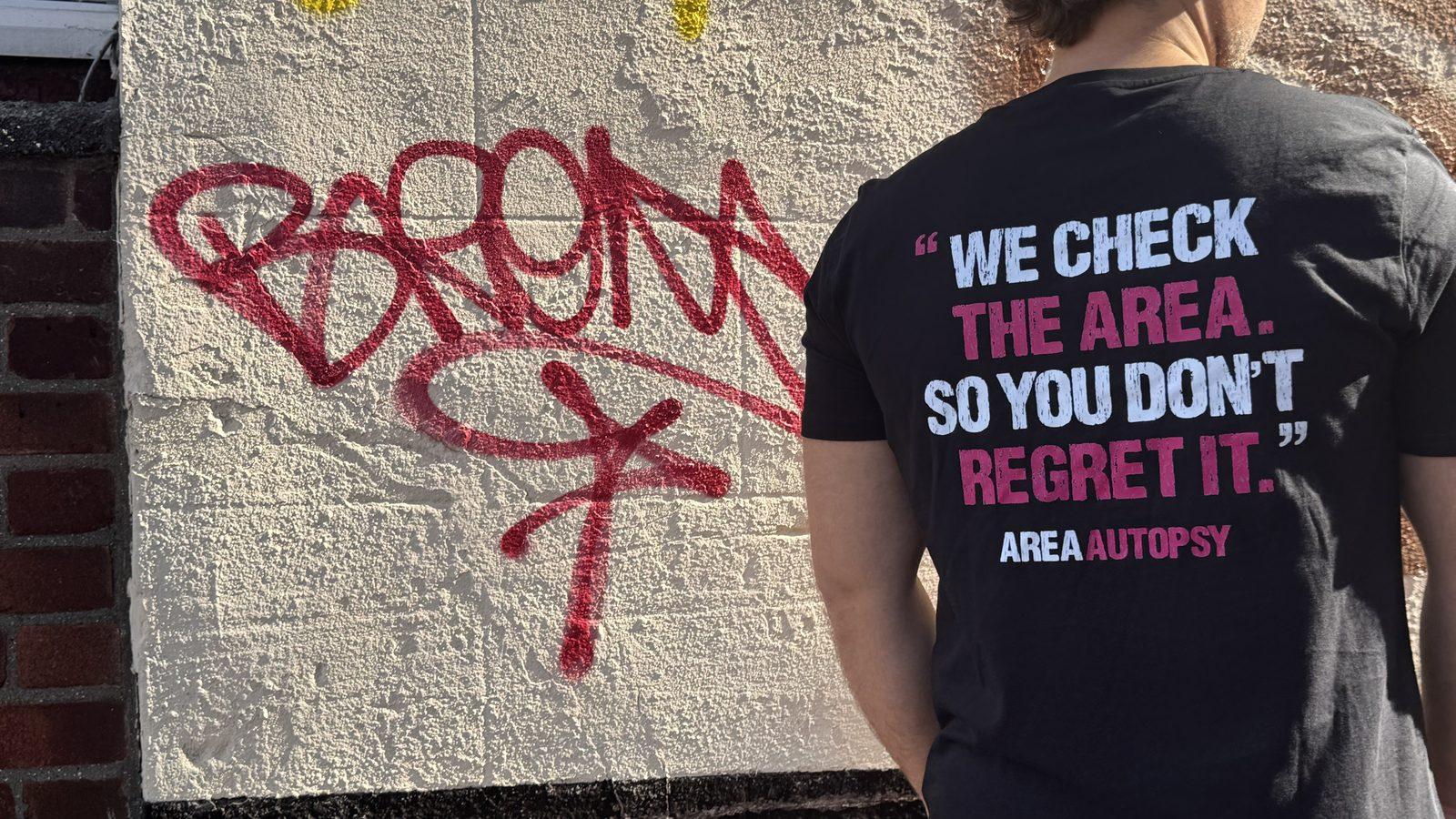 The back of an Area Autopsy t-shirt against a graffitied wall, reading “We check the area. So you don’t regret it.”